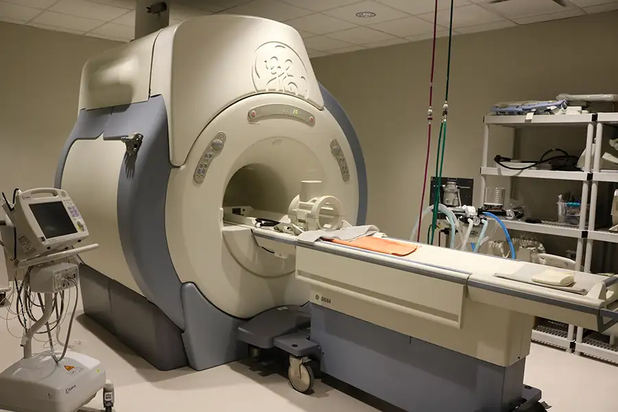 An MRI machine in a clinical setting, featuring a large, cylindrical scanner with a patient table extended in front. Medical equipment and monitors are visible to the side.