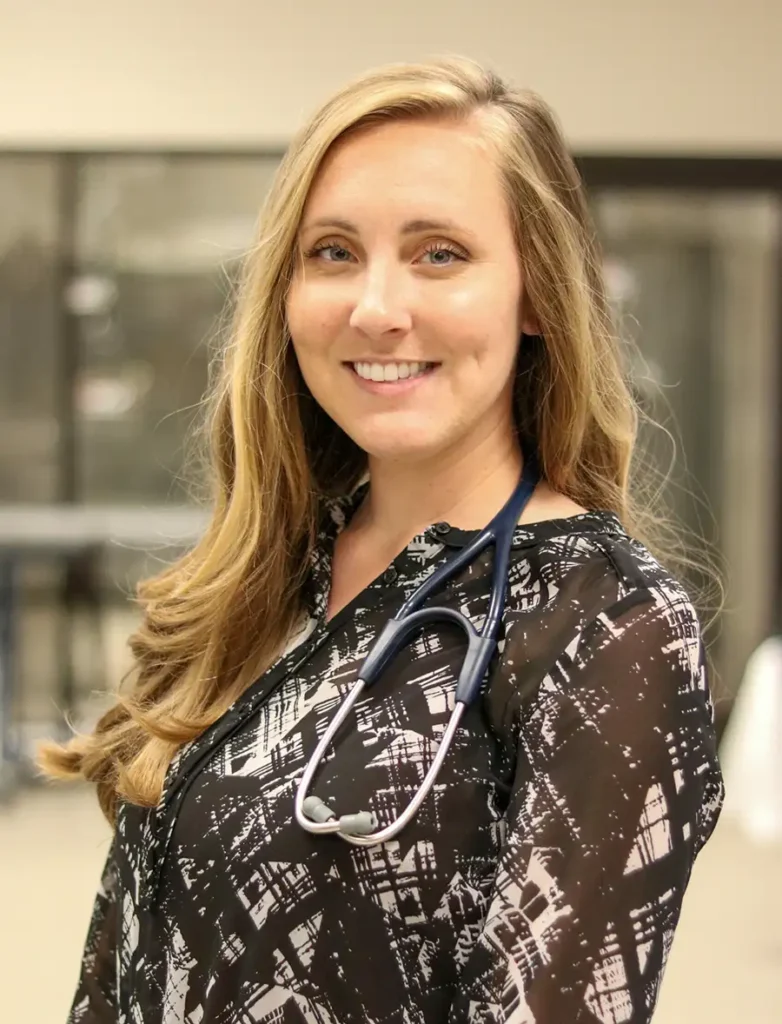 Portrait of Kim Cook smiling, wearing a black and white patterned blouse with a stethoscope around her neck in a professional medical setting.