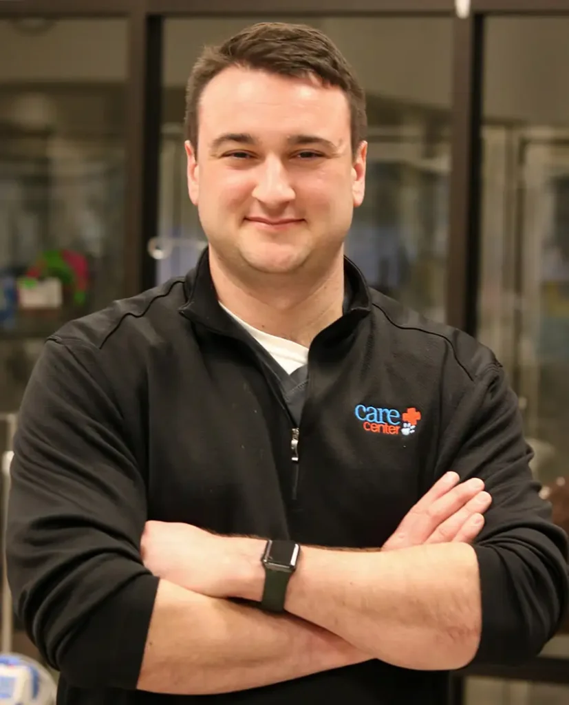 Portrait of Alec Repasy smiling with his arms crossed, wearing a black pullover with the 'Care Center' logo, in a professional medical setting.