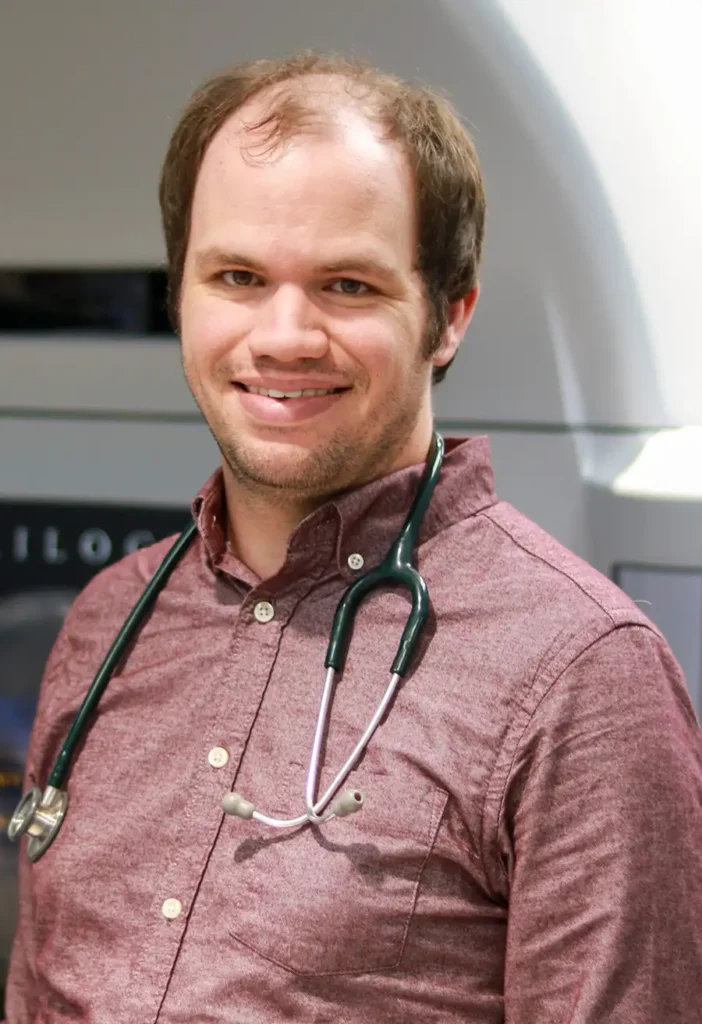 Portrait of Chris Bloom, smiling and wearing a maroon button-up shirt with a stethoscope around his neck, in a professional medical setting.