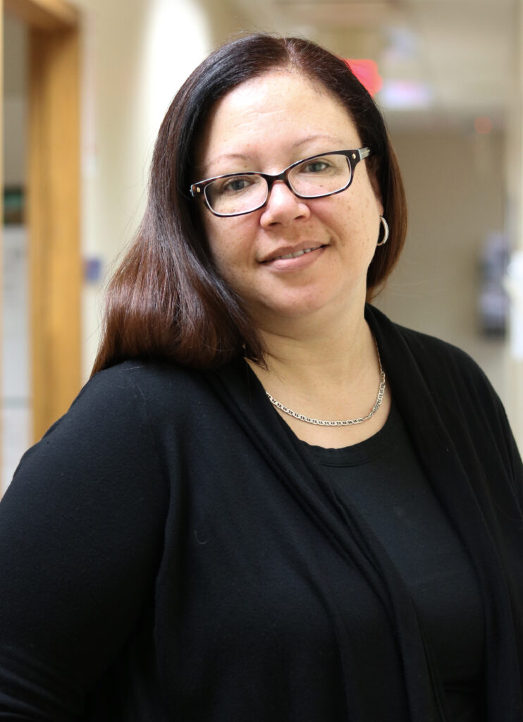 Portrait of Emily Stanley smiling, wearing a black top and black cardigan, with glasses and silver hoop earrings, with long auburn hair, in a professional setting.